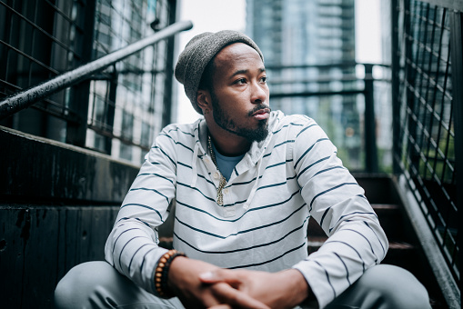 A Millennial  African American young adult man with casual street style and a beanie hat sits on some stairs in a downtown city scene.  Style, fashion, identity and youth culture in Seattle, Washington, USA.