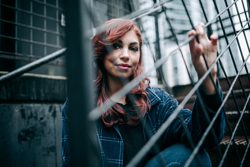 A Millennial multiracial young woman with casual street style looks through iron fence lattice in a downtown city scene.  Style, fashion, identity and youth culture in Seattle, Washington, USA.