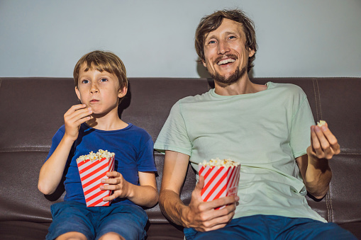 Father and son watching TV and eat popcort in evening at home.