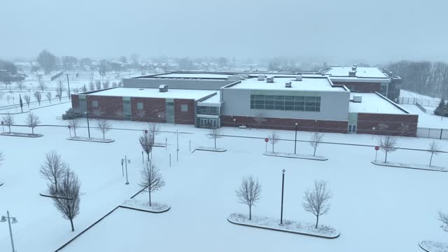 Closed school building in United States during snow day. Aerial orbit above snow-covered campus. Snow flurries falling. Public school cancellation theme.