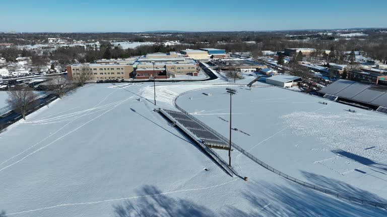 Aerial shot of a high school sports field covered in snow. Football stadium and track with bleacher seating. Winter in America theme.