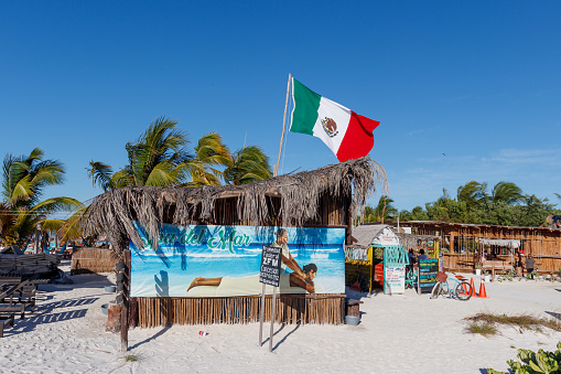 Holbox Mexico 12 04 2023: Flag of Mexico over blue sky with little hut and rancho and palm trees.