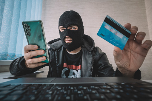 Male hacker in a robber mask uses phone, credit card and laptop in some fraudulent scheme. Cyber thief stole the personal data and credit card information. Hacker uses malware to steal user's money.
