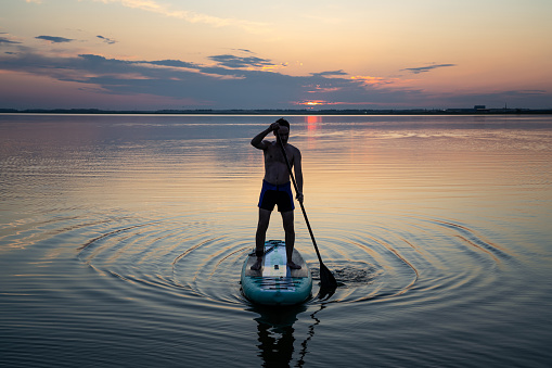 Standup paddleboarding. Silhouette of a guy rowing on SUP board in the sea against the background of the rising sun.