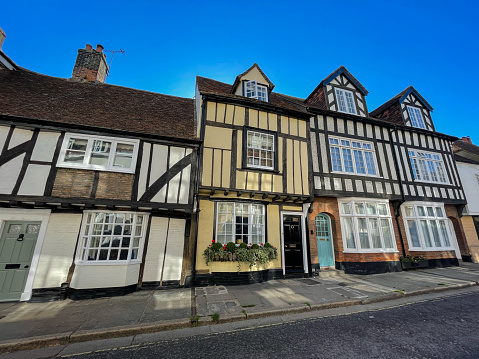 Tudor style homes with distinctive black and white timber framing under a clear blue sky, epitomizing classic British architecture. A picturesque old town street lined with historic terraced houses.