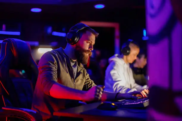 Bearded male eSports gamer playing online strategy video game with lots of action on a computer in a internet cafe or cybersport gaming lounge with her friends. Illuminated with neon lights.