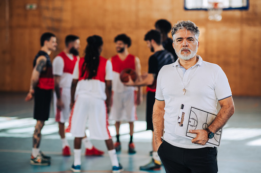 Portrait of senior basketball trainer standing on court with clipboard in his hands and looking at the camera while having training. In a blurry background are basketball players practicing basketball