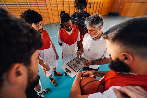 Senior basketball trainer is standing on court on training with his multicultural team with clipboard in hands and working on game strategy. An old coach is training his multiracial basketball team.