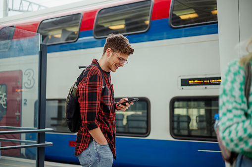 An urban traveler is standing at subway station and waiting for a metro while smiling at his phone. A trendy passenger is standing near the train and typing messages on his phone while smiling at it.