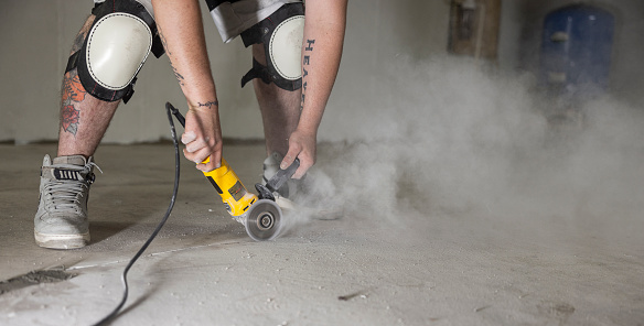 A construction worker is widening cracks in basement floor with angle grinder for waterproofing.