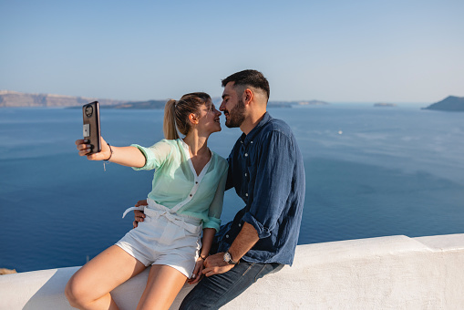 Cute young tourist couple in Santorini posing for a lovely selfie at a viewpoint. They are about to kiss. In The background, there is an amazing view of the coastline and the blue sea.