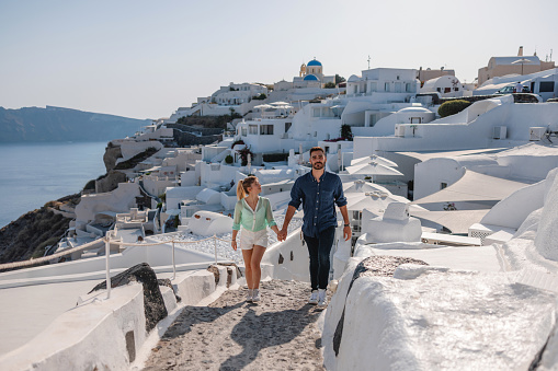 A young Caucasian tourist couple walking around the small alleys in Oia, Santorini. The attractive female and her boyfriend are holding hands as they explore a foreign country on a beautiful summer day.