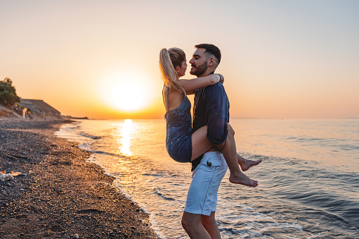 Romantic young adult Caucasian couple at the beach during sunset in Santorini. The boyfriend is holding his girlfriend up in the air while her legs are wrapped around him. They are looking at each other and smiling.