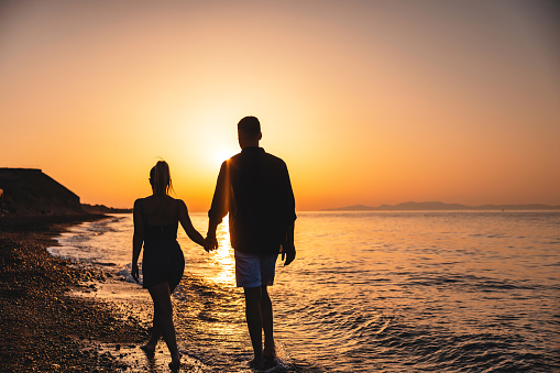 Back view of a romantic couple holding hands while walking towards the sunset at the beach. Only silhouettes are visible. The sky is orange. Romantic date on the beach in Santorini.