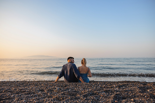 Back view of a romantic young tourist couple enjoying a beach date in Greece. They are sitting side by side with their arms around each other. The sun is setting and the sea is calm. Romantic moments on couple summer vacations.