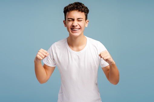 Excited strong teenager wearing stylish white t shirt rejoicing and supporting favourite team with closed eyes isolated on blue background. Advertisement concept