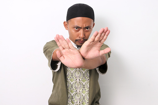 Indonesian Muslim man in koko shirt and peci crosses his arms in a stop gesture. Isolated on White background