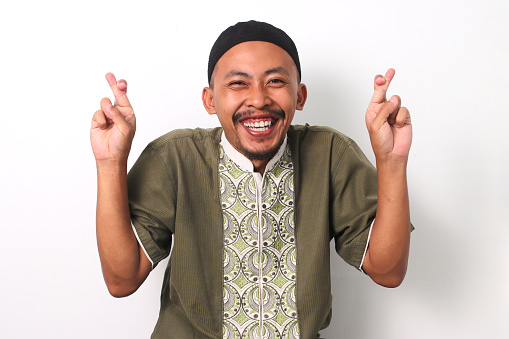 Indonesian Muslim man in koko shirt and peci celebrates with crossed fingers, expressing joy and good fortune. Isolated on White background