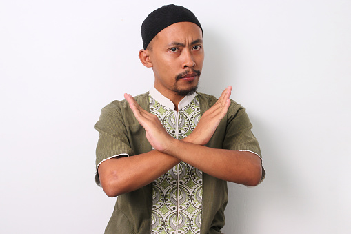 Indonesian Muslim man in koko shirt and peci crosses his arms in a stop gesture. Isolated on White background
