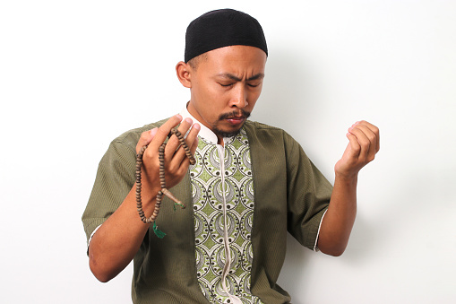 An Indonesian Muslim man in koko and peci prays with devotion during Ramadan, his open hands raised in supplication to Allah. Isolated on a white background