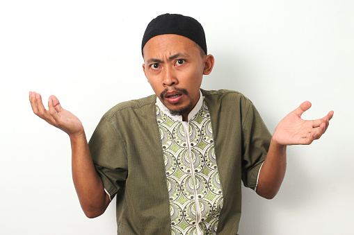 An Indonesian Muslim man in koko and peci shrugs his shoulders and raises his arms in a gesture of uncertainty or confusion. Isolated on a white background