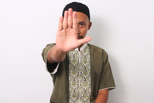 An Indonesian Muslim man in koko and peci makes a stop gesture, demonstrating his commitment to avoiding prohibitions during the holy month of Ramadan. Isolated on a white background