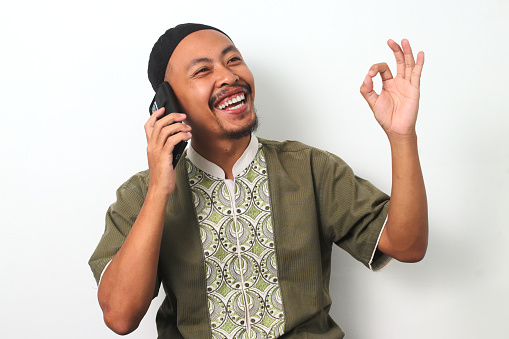 Happy Indonesian Muslim man in koko and peci makes an OK gesture while talking on his phone, expressing satisfaction or agreement during Ramadan. Isolated on a white background