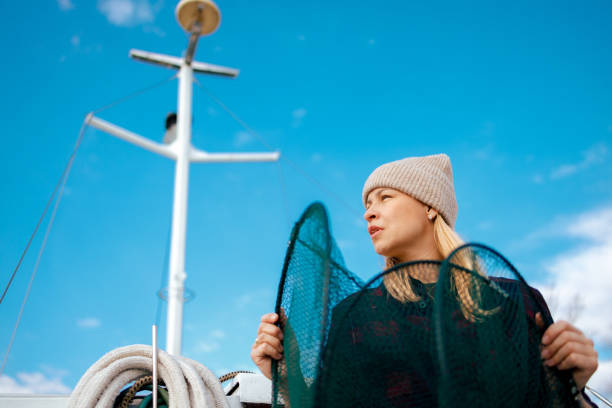 A woman in a warm beanie looks into the distance as she holds a green fisherman's net aboard a boat, with the clear blue sky above and the mast of the boat standing tall in the background.