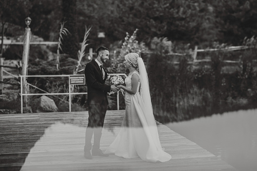 The groom in a black suit and white shirt gently touches and holds the hand of the bride in a white wedding dress, standing near a lake and stones. Black and white photo
