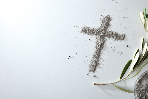 Cross made with ashes on white table with olive leaves and container full of ashes. Top view.