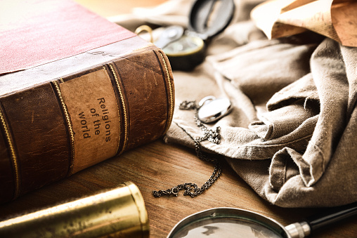 Old and deteriorated book of the religions of the world with ancient objects of study and adventure on wooden table. Elevated view.