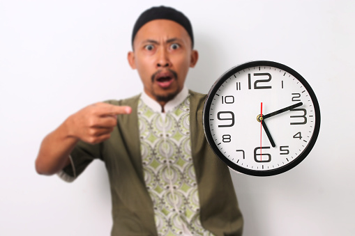 A shocked Indonesian Muslim man in koko and peci emphatically points at a clock, realizing he's late for his sahur meal during Ramadan. Isolated on a white background