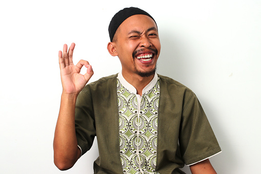 Happy Indonesian Muslim man in koko and peci gives a playful wink and an OK gesture, expressing satisfaction and positive vibes during Ramadan. Isolated on a white background.
