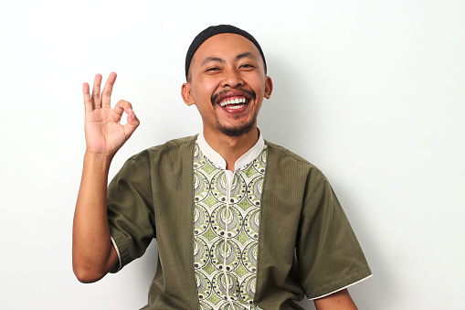 Happy Indonesian Muslim man in koko and peci shows an OK gesture, expressing satisfaction and positivity during Ramadan. Isolated on a white background