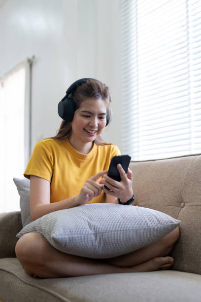 young asian woman using mobile phone and earphones to listening to music or online chatting at home - room service audio 뉴스 사진 이미지