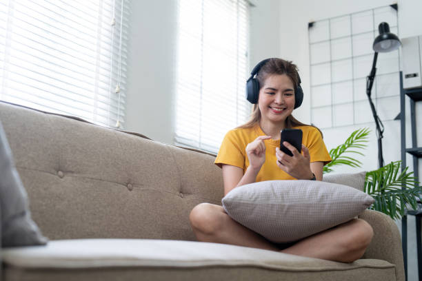 young asian woman using mobile phone and earphones to listening to music or online chatting at home - room service audio стоковые фото и изображения