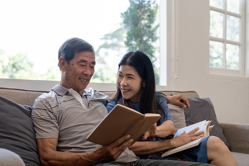 Asian senior couple reading book diary together, sitting on cozy sofa in home interior. Happy retirement activity lifestyle at home.