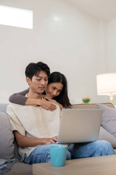 istock Happy asian couple using laptop sit relax on sofa doing ecommerce shopping online on website at home 2049445906
