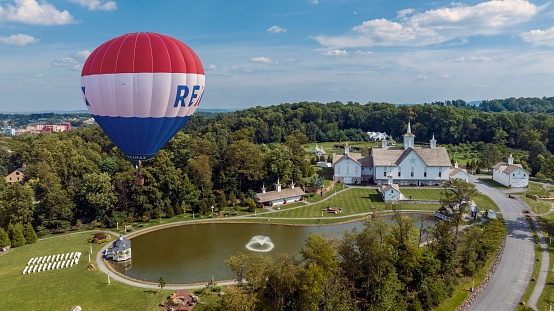 Elizabethtown, Pennsylvania, USA, August 8, 2023 - Vibrant Hot Air Balloon With Red, White, And Blue Stripes And Large Letters Floating In The Sky Above A Scenic Landscape Buildings, A Reflective Pond