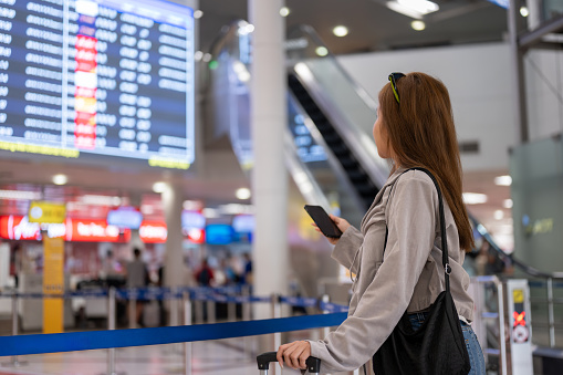 An attractive Asian female traveler is standing in front of a boarding time monitor in the airport, checking her flight boarding time. people and transportation concepts