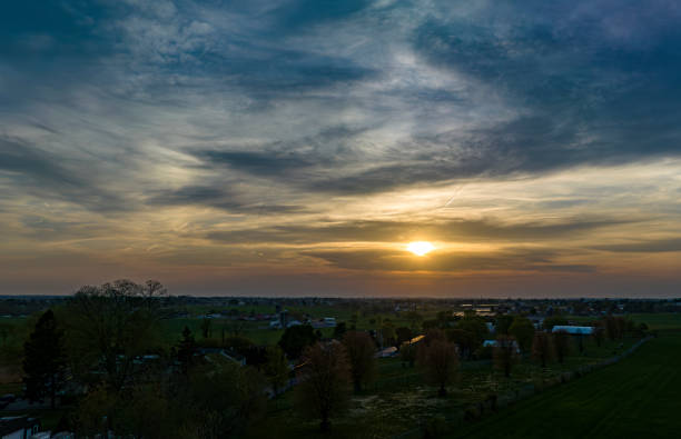 Dramatic Sunset Casting A Warm Glow Over A Rural Landscape With Silhouetted Trees And Buildings Under A Cloud-Streaked Sky.