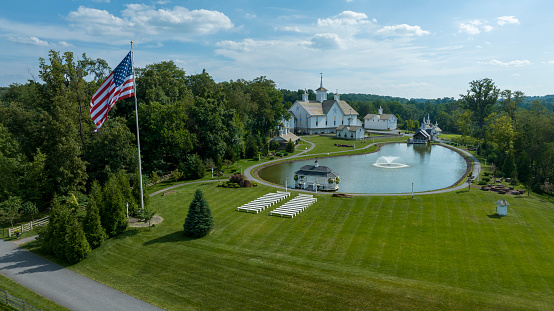 Aerial View Of A Grand White Building With A Grey Roof Surrounded By Lush Green Lawns, A Large Reflective Pond With Fountains, And A Majestic Flagpole Bearing The American Flag.