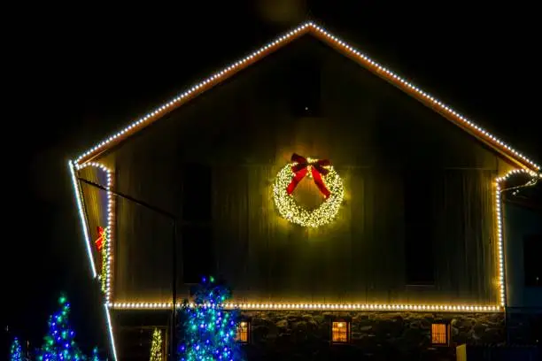 Close-Up Night Image Of A Building's Gable Trimmed With White Christmas Lights And A Large Lit Wreath, With Blue-Lit Trees In The Foreground.