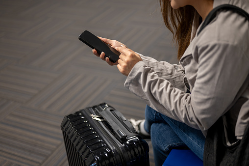 A close-up shot of a beautiful Asian female traveler passenger is using her smartphone while sitting at a waiting seat in the airport, waiting for her boarding time.