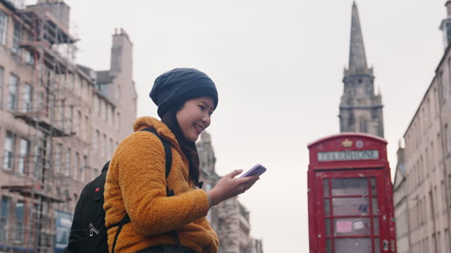 Asian woman pedestrian traveler wearing winter coat using mobile phone and smiling with red public telephone booths and looking around in Old Town, Edinburgh, Scotland, United Kingdom