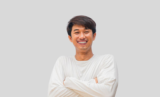 A young Asian man in his 20s wearing a white t-shirt stands confidently with his arms crossed over his chest, isolated on a gray background.