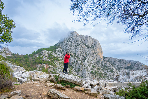 A man with a red jacket and long hair takes photos of the ancient city of Termessos with a camera. He is standing still. Gulluk Mountain, Antalya
