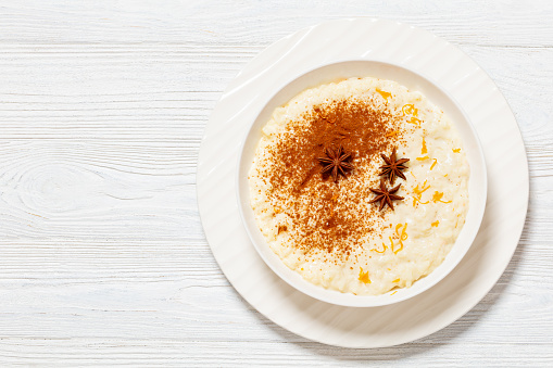 rizogalo, creamy greek rice pudding of whole milk, white rice, cinnamon, orange and lemon zest in white bowl on white wooden table, horizontal view from above, flat lay, free space