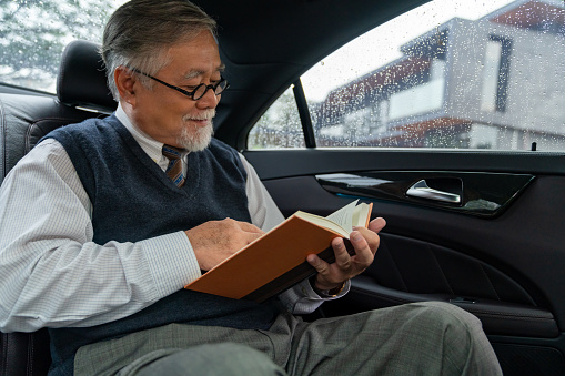Confidence senior businessman CEO in suit wearing glasses sitting on car back seat and writing business plan in the book while going to work at office. Elderly businessman and transportation concept.
