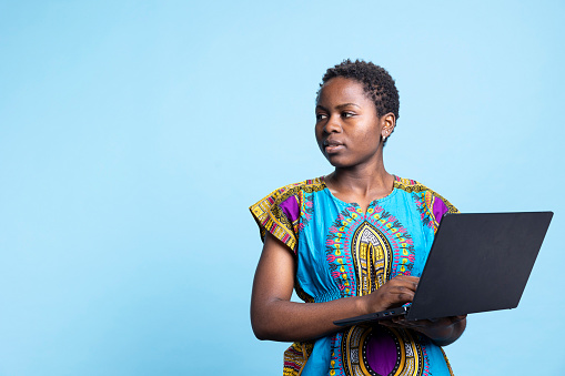 African American woman using wireless connection on pc in front of camera to access websites and social networks. Young person uses a web link to enter data digitally, blue background studio.